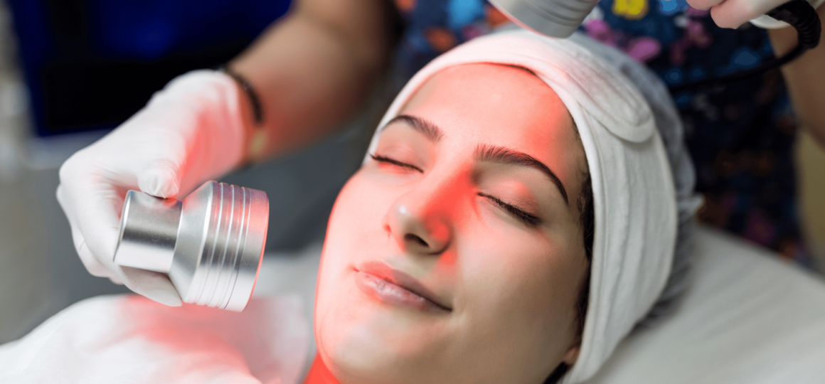 A woman with closed eyes receives red light therapy for acne as she lies on a table in a spa-like setting.